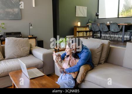 Couple sitting on couch video chatting with friends on laptop at home Stock Photo