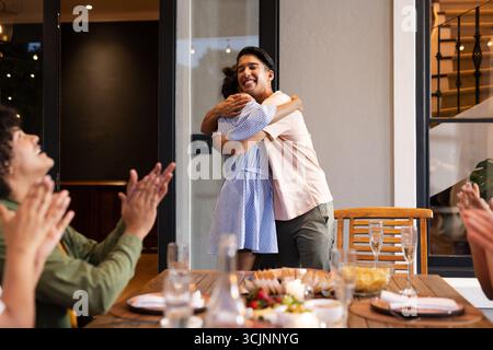 Diverse friends celebrating at dinner party, young couple hugging while others clapping Stock Photo