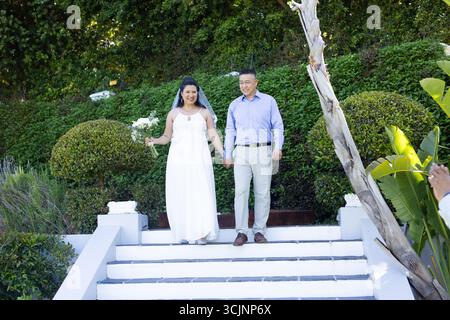 Walking down garden steps, bride and groom holding hands smiling joyfully Stock Photo