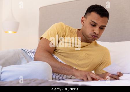 Young man lying on bed looking at photograph, appearing thoughtful and relaxed Stock Photo