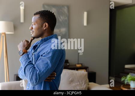 Thoughtful man in blue shirt contemplating at home, looking out window Stock Photo