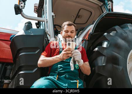 Man farmer checking his smartphone near tractor outdoors in countryside. Agricultural mobile technology. Stock Photo