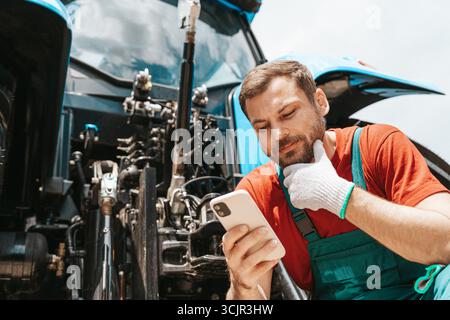 Modern farmer using digital technology in agriculture. Man using smartphone near tractor. Stock Photo