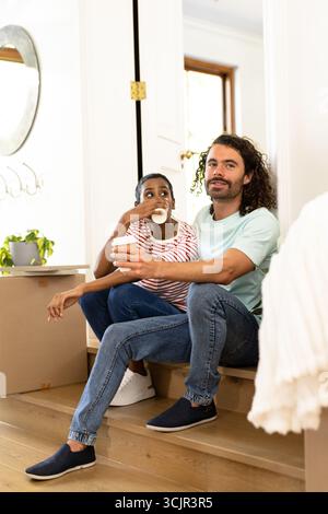 Young diverse couple sitting on stairs at home, enjoying coffee and relaxing together Stock Photo