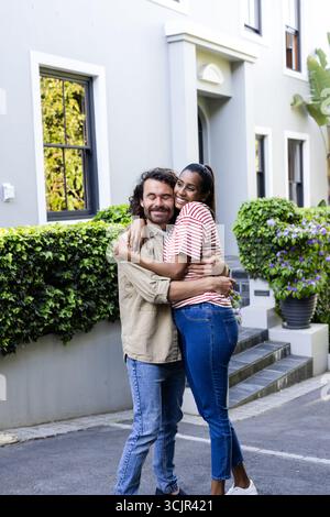 Diverse couple embracing happily outside modern home, enjoying sunny day together Stock Photo