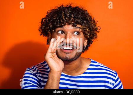 Cheerful young man in a striped shirt making a playful gesture against a bright orange background, expressing joy Stock Photo