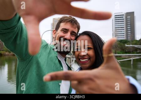Happy multiracial couple taking a photo framing with hands in a city park Stock Photo