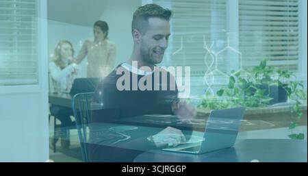 Male staff typing on laptop at round table in office lounge, with potted plants and blinds Stock Photo