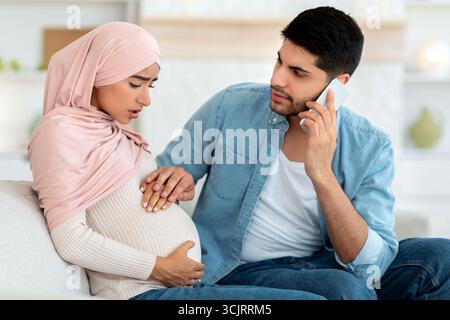 Pregnant arab woman having prenatal contractions, worried husband calling doctor, using mobile phone, home interior Stock Photo