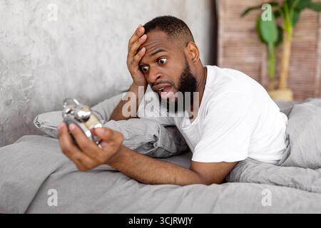 Shocked black man looking at clock in bed Stock Photo