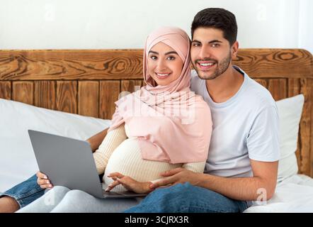 Loving pregnant muslim couple using laptop together, surfing internet and spending time together, sitting on bed Stock Photo