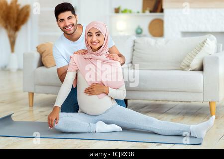 Pregnancy workout. Happy arab spouses exercising at home, woman stretching legs while man massaging wife's shoulders Stock Photo