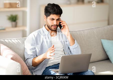 Computer problems. Displeased arab man talking on smartphone and looking on laptop screen at home Stock Photo