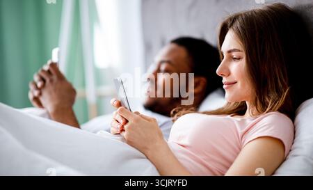 Gadget addiction concept. Young multiethnic family using smartphones while lying in bed, copy space Stock Photo
