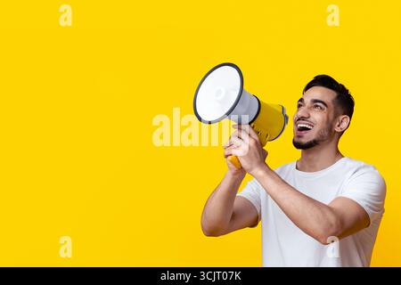 Excited arab guy speaking loud, using megaphone on yellow Stock Photo
