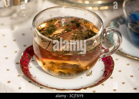 Glass cup filled with brown hot herbal tea, infused with dried flowers and herbs, placed on a decorative saucer. A warm, cozy beverage Stock Photo
