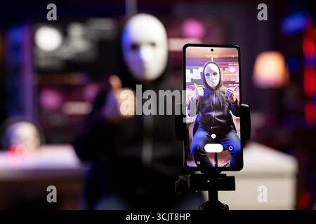Close up of masked evil man using mobile phone to film ransom video in secret base. Focus on smartphone used by scammer wearing anonymous mask to record blackmail threats in neon apartment Stock Photo