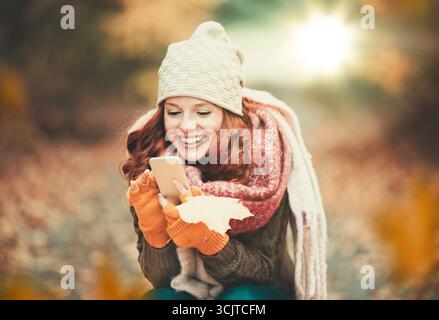 red-haired cheerful woman in autumn looking at her smartphone Stock Photo