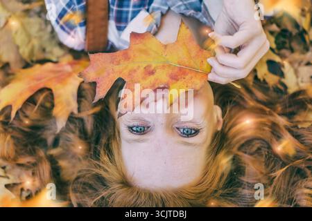 red-haired woman lying in autumn leaves, holding an autumn leaf in front of her mouth, portrait Stock Photo