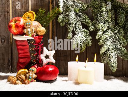 filled Santa's boot, candles and fir branch in front of a wooden wall as Christmas decoration Stock Photo