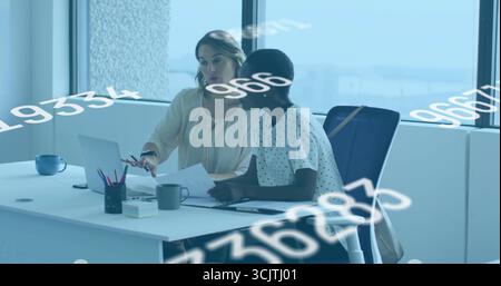 Reviewing two women wearing business attire collaborating at desk near window, with silver laptop Stock Photo