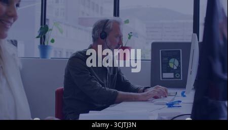Typing senior worker wearing headset and dark shirt at office desk, with computer displaying charts Stock Photo