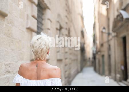 A woman in a white dress walks down a narrow alleyway. The alleyway is lined with buildings, and the woman is looking up at something. The scene has a Stock Photo