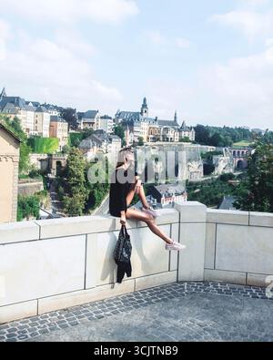 Fashion model is sitting and enjoying spectacular city view of old city of Luxembourg, while traveling. Beautiful woman exploring Stock Photo