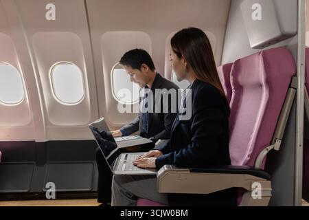 A business man and woman in formal suits working together on a flight. They are discussing documents and using laptops, showing teamwork and productiv Stock Photo