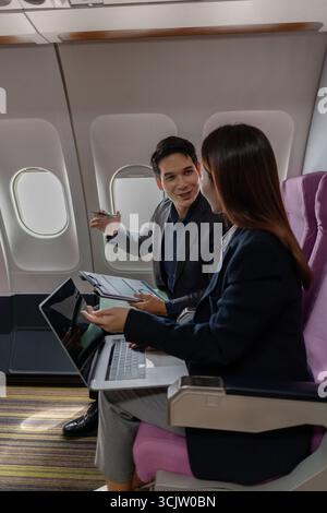A business man and woman in formal suits working together on a flight. They are discussing documents and using laptops, showing teamwork and productiv Stock Photo