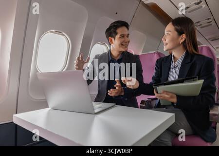 A business man and woman in formal suits working together on a flight. They are discussing documents and using laptops, showing teamwork and productiv Stock Photo