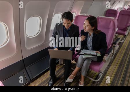 A business man and woman in formal suits working together on a flight. They are discussing documents and using laptops, showing teamwork and productiv Stock Photo