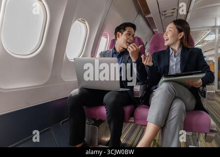 A business man and woman in formal suits working together on a flight. They are discussing documents and using laptops, showing teamwork and productiv Stock Photo