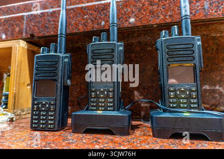 Three walkie talkies sit on charging docks with indicator lights on, ready for communication and emergency response use. Stock Photo