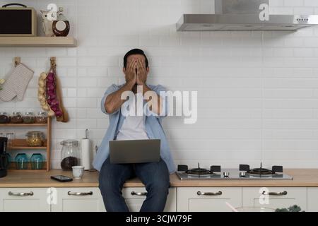 Upset young man sitting on kitchen counter covering face with hands in frustration while using laptop, concept of stress, failure, remote work problem Stock Photo