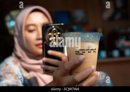 Young woman in hijab captures iced coffee with her phone in a cafe setting, showcasing a modern and trendy lifestyle image perfect for social media us Stock Photo