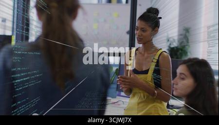Presenting woman in yellow overalls explaining code in office workstation, sticky-note whiteboard Stock Photo