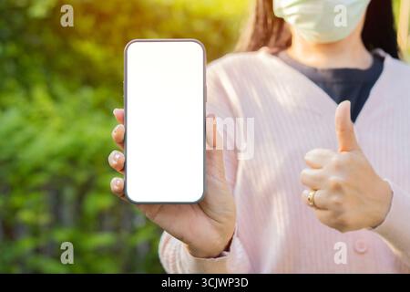 Online service, technology. Close-up of mobile phone with isolate screen holding by woman wearing medical mask with show thumb up. Stock Photo
