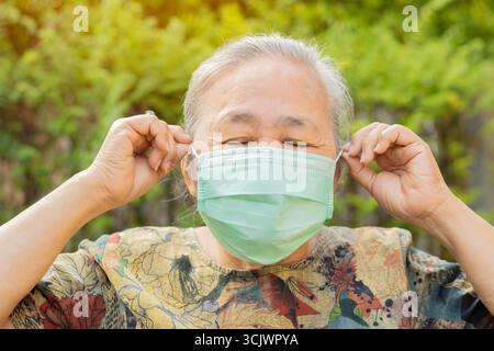 Portrait of female senior wearing medical protective mask and close her eyes while standing against nature background. Stock Photo