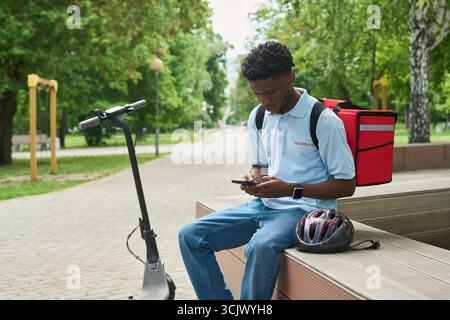 Black Man Using Smartphone While Sitting Outdoors with Electric Scooter Stock Photo