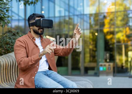 Young man is interacting with virtual reality using a vr headset outdoors, exploring immersive technology against a backdrop of modern architecture Stock Photo