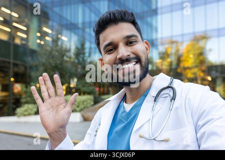 Portrait of a smiling indian doctor waving hand in front of a modern hospital building, radiating friendliness and approachability while showcasing confidence and professionalism Stock Photo