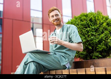 Smiling man in scrubs holding takeaway drink while using laptop Stock Photo