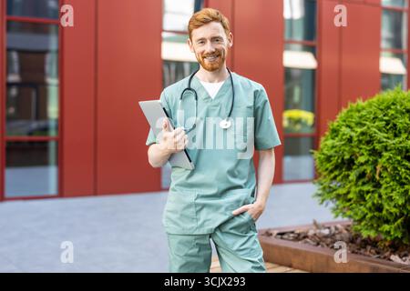 Smiling man doctor in scrubs standing outdoors holding closed laptop Stock Photo