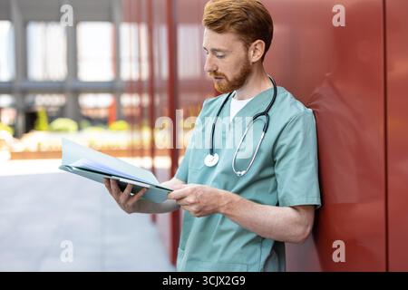 Man in green uniform leaning on wall with open patient records Stock Photo