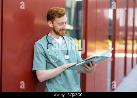 Man doctor standing by wall reading from open medical folder Stock Photo