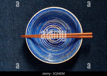A pair of wooden chopsticks lying across an empty soup bowl on blue fabric background, close up, top view Stock Photo