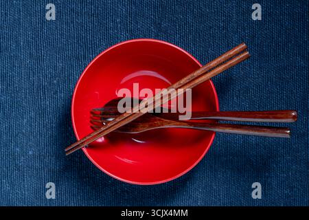 A pair of wooden chopsticks, fork and spoon lying across an empty red soup bowl on blue fabric background, close up, top view Stock Photo