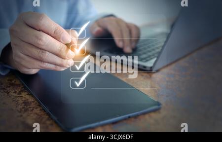 Hand checking digital checklist on tablet with computer, illustrating task organization, corporate efficiency, and productivity strategy for business Stock Photo