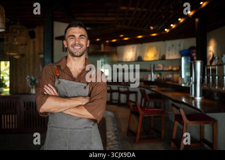 Proud cafe owner standing confidently behind the counter, ready to serve customers with a warm smile Stock Photo
